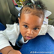 Aloura is registered to the contest to win money with this photo: baby, child, infant, portrait, car, car_interior, seated, close_up, face, eyes, curly_hair, hair_buns, hair_ties, white_shirt, blue_clothing, hand, indoor, passenger, looking_at_camera, cute