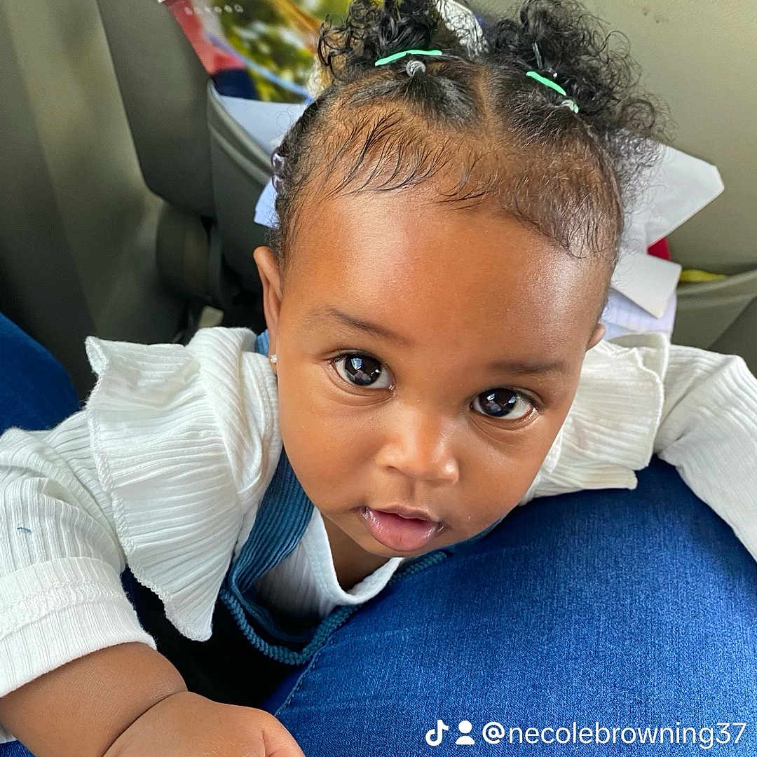 Aloura is registered to the contest to win money with this photo: baby, child, infant, portrait, car, car_interior, seated, close_up, face, eyes, curly_hair, hair_buns, hair_ties, white_shirt, blue_clothing, hand, indoor, passenger, looking_at_camera, cute