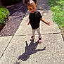 child, outdoor, sidewalk, shadow, butterfly_hair_clips, curly_hair, black_shirt, leggings, sneakers, sunlight, greenery, brick_wall, cute, fashion, standing, posing, daytime, playful, confident, person