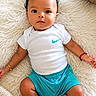 baby, child, curly_hair, white_onesie, nike_logo, teal_shorts, bracelets, fluffy_rug, cream_rug, indoor, portrait, cute, infant, lying_down, looking_at_camera, skin, footwear_absent, relaxed, decor, plant
