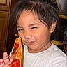 battery_icon, cabinet, camera_icon, child, clutter, face, flashlight_icon, food, hair, hand, holding, indoor, pepperoni, person, phone_screen, pizza, smile, squinting, time_display, white_tshirt