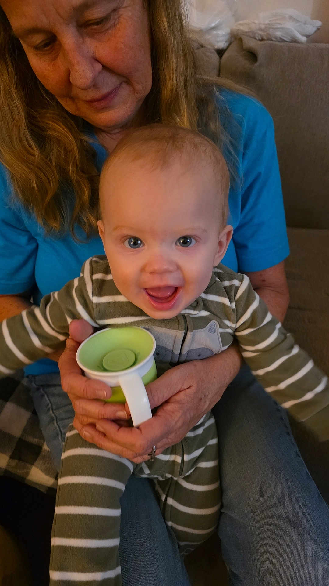 Branch is registered to the contest to win money with this photo: baby, child, woman, adult, holding, sippy_cup, striped_clothing, pajamas, blue_shirt, couch, blanket, indoor, smiling, happy, person, face, hands, seated, home, casual