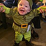Branch is registered to the contest to win money with this photo: baby, child, costume, green_clothing, hat, smiling, indoor, floor, person, happy, standing, hands, adult, supporting, footwear, wooden_floor, home, cute, red_bow, playful
