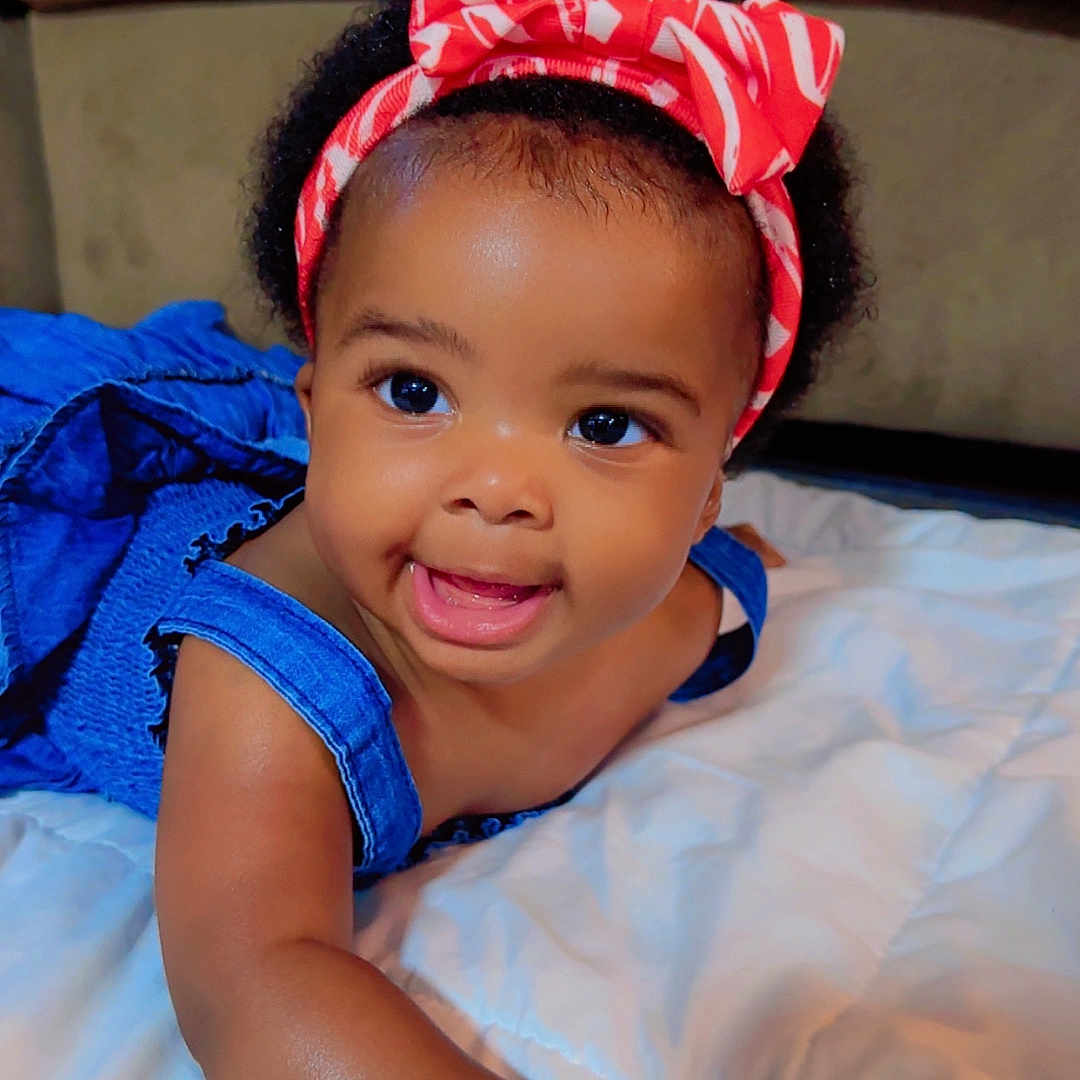 Reina is registered to the contest to win money with this photo: baby, black_hair, blanket, blue_dress, bright_eyes, child, couch, cute, face, happy, headband, indoors, infant, joyful, lying_down, person, portrait, skin, smiling, toddler
