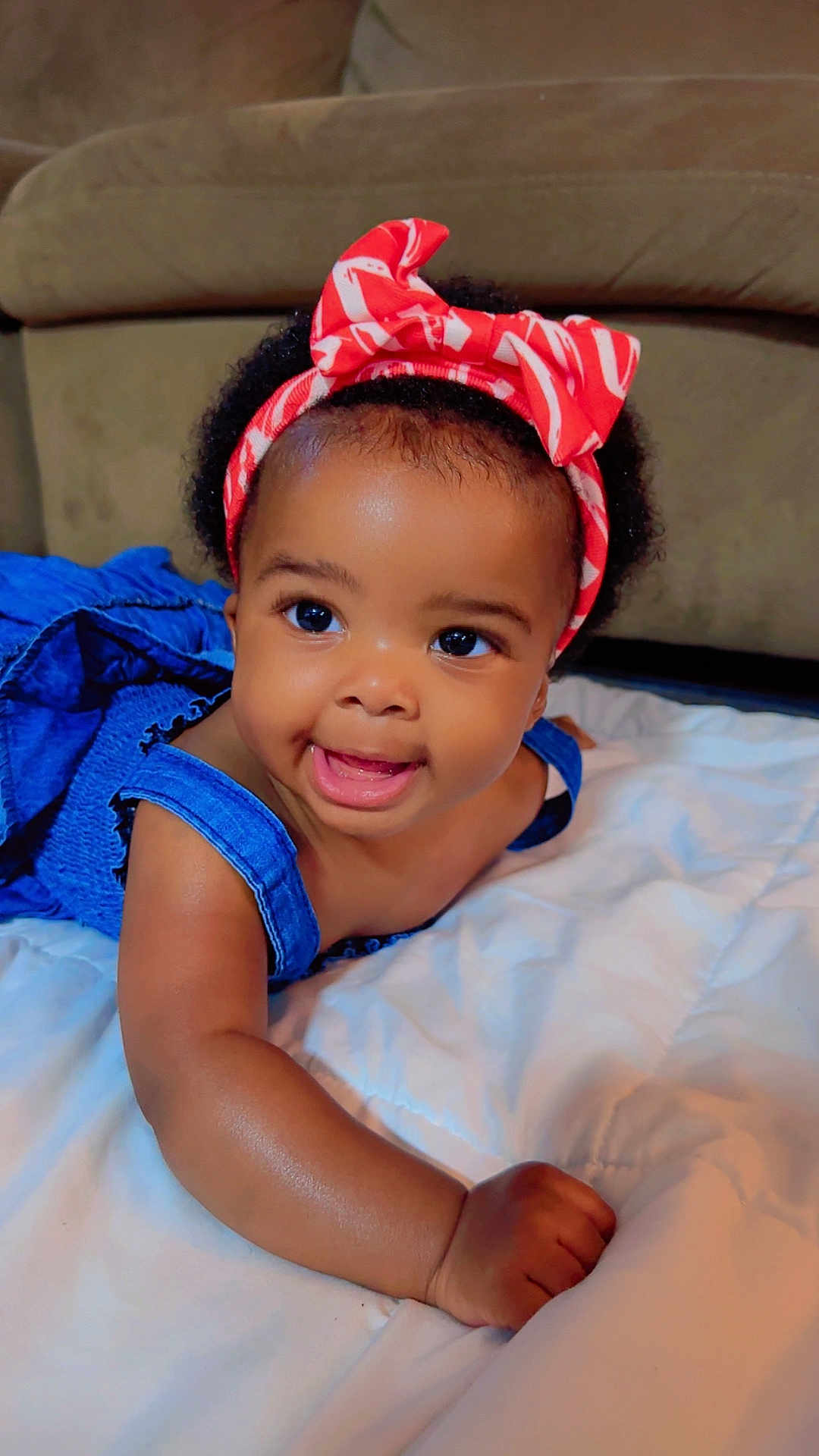 Reina is registered to the contest to win money with this photo: baby, child, smiling, headband, blue_dress, cute, lying_down, blanket, indoors, happy, infant, skin, face, person, portrait, couch, black_hair, bright_eyes, joyful, toddler