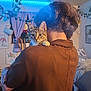 cat, person, indoor, brown_sweater, tabby_cat, curious, shoulder, plant, curtain, framed_picture, blue_led_light, home_decor, bed, blanket, wooden_floor, hand, hair, window, couch, lighting