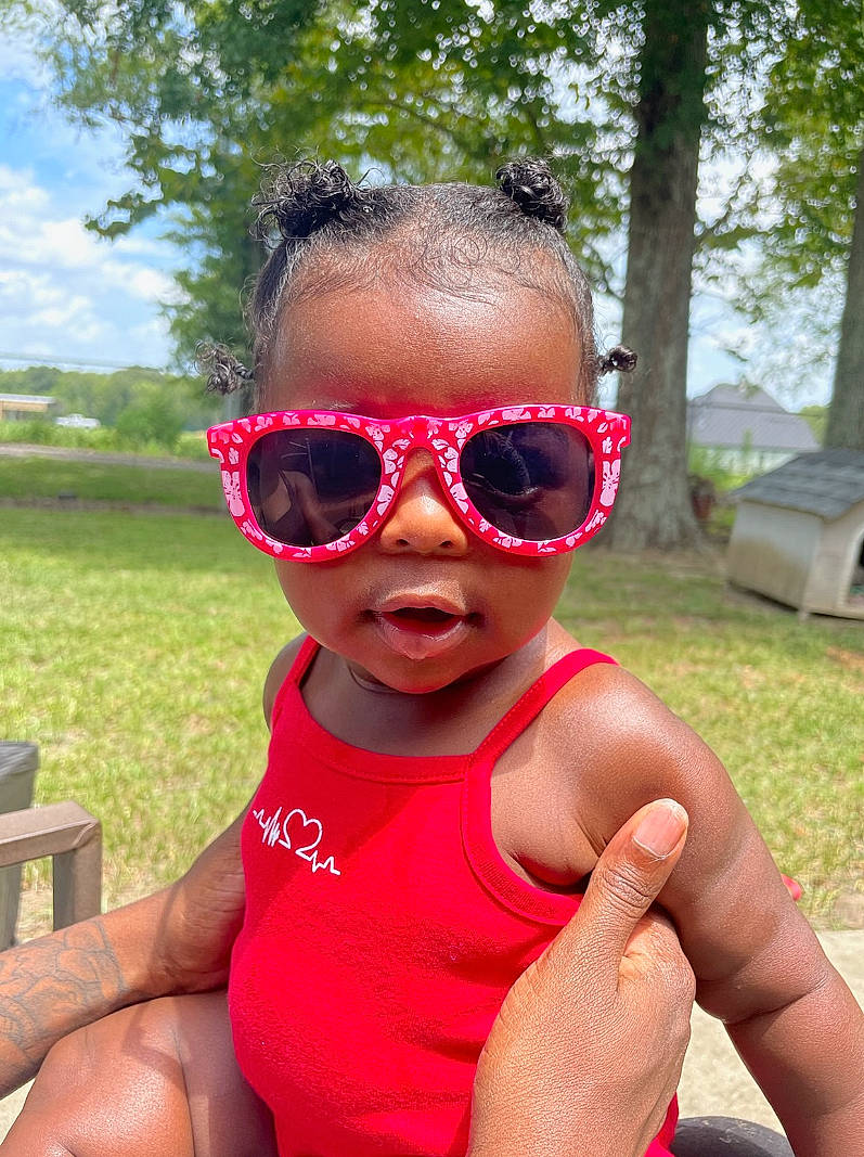 Rihanna is registered to the contest to win money with this photo: baby_toddler_clothing, eyewear, goggles, grass, happy, leaf, leisure, lip, people_in_nature, person, personal_protective_equipment, pink, plant, recreation, red, sky, summer, sunglasses, toddler, tree