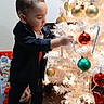 child, toddler, christmas_tree, ornaments, candy_canes, holiday, decorations, indoor, curious, black_clothing, shoes, floor, presents, festive, white_tree, lights, standing, holiday_season, celebration, home