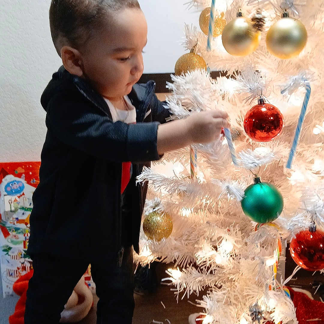 Carinlefty is registered to the contest to win money with this photo: child, toddler, christmas_tree, ornaments, candy_canes, holiday, decorations, indoor, curious, black_clothing, shoes, floor, presents, festive, white_tree, lights, standing, holiday_season, celebration, home