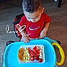 baby, casual_clothing, child, colorful, curious, cute, floor, food, furniture, indoor, meal, person, plate, red_shirt, text_overlay, toddler, toy, walker, wooden_floor, young_child