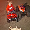 asphalt, blurred_motion, casual_clothing, child, honda, looking_sideways, night, nike, outdoor, parking_lot, person, playing, red_clothing, sitting, small_vehicle, street, toy_car, toy_vehicle, wheels, young_child