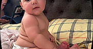 Arya is registered to the contest to win money with this photo: baby, child, diaper, plaid_bedspread, bed, headboard, curly_hair, chubby, skin, bare_skin, indoor, person, sitting, looking, big_eyes, hand, foot, adult, phone, cozy