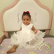 Inaya participe au concours pour gagner de l'argent avec cette photo : toddler, child, dress, white_dress, bow, bed, headboard, unicorn, blanket, smiling, indoors, furniture, colorful, happy, person, cute, hair_bow, sitting, foot, playful
