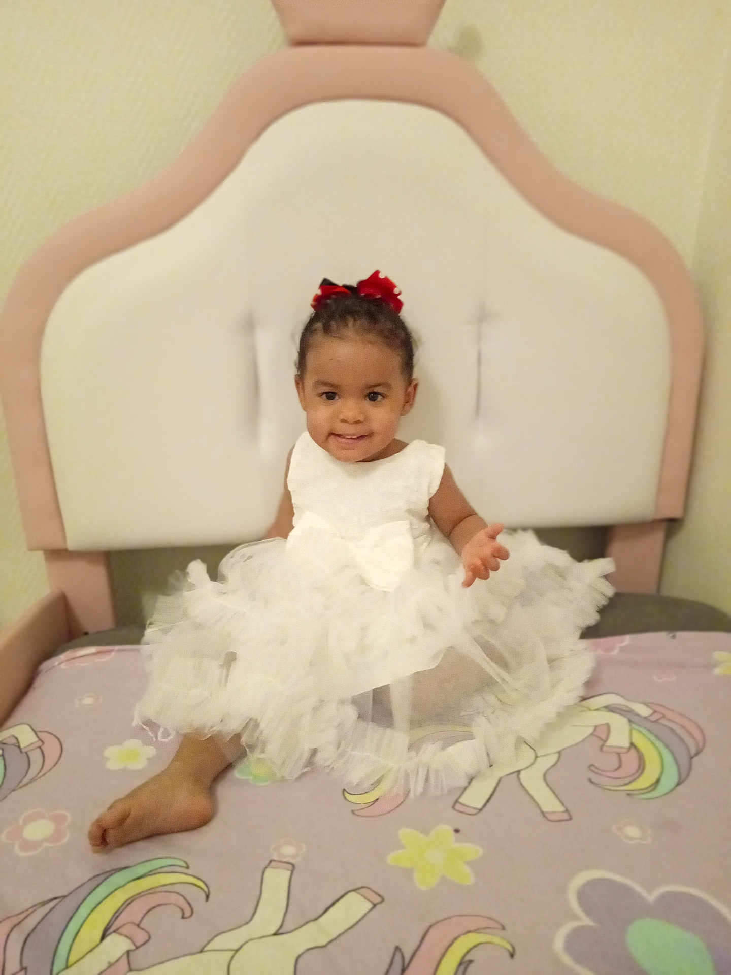Inaya participe au concours pour gagner de l'argent avec cette photo : toddler, child, dress, white_dress, bow, bed, headboard, unicorn, blanket, smiling, indoors, furniture, colorful, happy, person, cute, hair_bow, sitting, foot, playful