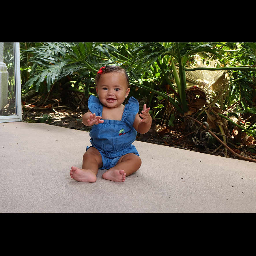 Jaliana Collins joined the competition — help win amazing prizes! baby, blue_clothing, child, cute, face, feet, greenery, hands, happy, infant, nature, outdoor, patio, plants, red_bow, romper, sitting, skin, smiling, sunlight