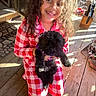 child, dog, puppy, pajamas, red_checkered, curly_hair, smiling, indoors, wooden_floor, sunlight, kneeling, pet, happy, casual, cozy, home, black_dog, young, cute, holding