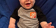 Santiago is registered to the contest to win money with this photo: baby, infant, smiling, sitting, blue_cushion, clothing, shirt, home_depot_logo, hand, face, eyes, happy, cute, child, person, portrait, indoors, cozy, young, skin