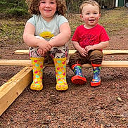 Bodie is registered to the contest to win money with this photo: child, children, outdoor, forest, boots, rain_boots, wood, smile, happy, nature, grass, trees, play, sitting, casual_clothes, toddler, cute, daylight, two_people, ground