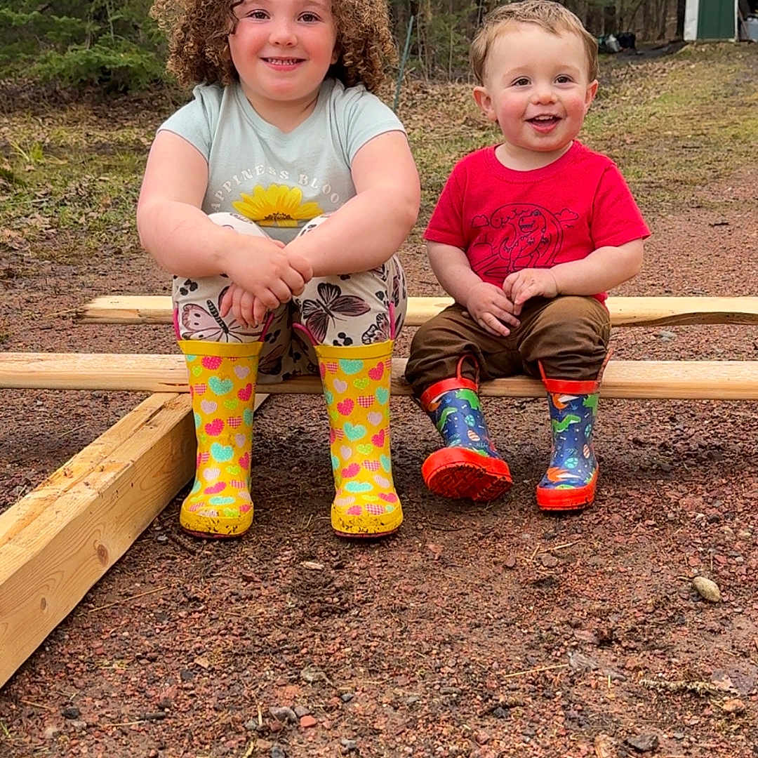 Bodie is registered to the contest to win money with this photo: child, children, outdoor, forest, boots, rain_boots, wood, smile, happy, nature, grass, trees, play, sitting, casual_clothes, toddler, cute, daylight, two_people, ground