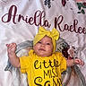 Ariella is registered to the contest to win money with this photo: baby, infant, yellow_clothing, headband, floral_blanket, text, lying_down, cute, child, portrait, indoors, person, young_child, smiling, bright_colors, soft_texture, happy, face, hand, apparel