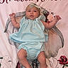 baby, infant, blue_outfit, headband, blanket, floral_pattern, name_text, lying_down, cute, small_hands, small_feet, newborn, child, portrait, indoors, pink_background, soft_texture, baby_clothes, looking_up, adorable