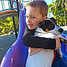 child, dog, hugging, slide, playground, outdoor, sunny, trees, blue_sky, smiling, animal, pet, boy, leash, park, daytime, happy, casual_clothing, fun, nature