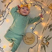 Livio a rejoint le concours — aidez-le/la à gagner de superbes lots ! baby, crown, fairy_lights, green_pajamas, crib, animal_print, giraffe, wooden_plaque, milestone, two_months, smiling, soft_blanket, cozy, infant, cute, decorative_lights, cushions, lying_down, indoors, child