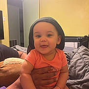 Jaelynn joined the competition — help win amazing prizes! baby, child, infant, smiling, orange_onesie, black_cap, hand, indoor, bed, blanket, toy, playful, person, face, cute, happy, holding, room, casual, family