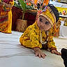 child, baby, toddler, crawling, traditional_clothing, yellow_outfit, headband, jewelry, necklace, face_paint, festival, indian_culture, religious_shrine, decorations, flowers, plant, indoor, tile_floor, portrait, colorful