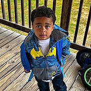 Kaynan is registered to the contest to win money with this photo: child, boy, hoodie, batman, blue, yellow, backpack, wooden_deck, outdoor, grass, bicycle, curious, standing, casual_clothing, young, person, footwear, daylight, portrait, expression