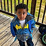child, boy, hoodie, batman, blue, yellow, backpack, wooden_deck, outdoor, grass, bicycle, curious, standing, casual_clothing, young, person, footwear, daylight, portrait, expression