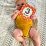 baby, child, chubby_legs, cute, eyes, face, giraffe_print, hand_holding_card, headband, indoors, infant, lying_down, milestone_card, orange_card, patterned_blanket, portrait, quilt, skin, soft_lighting, yellow_onesie