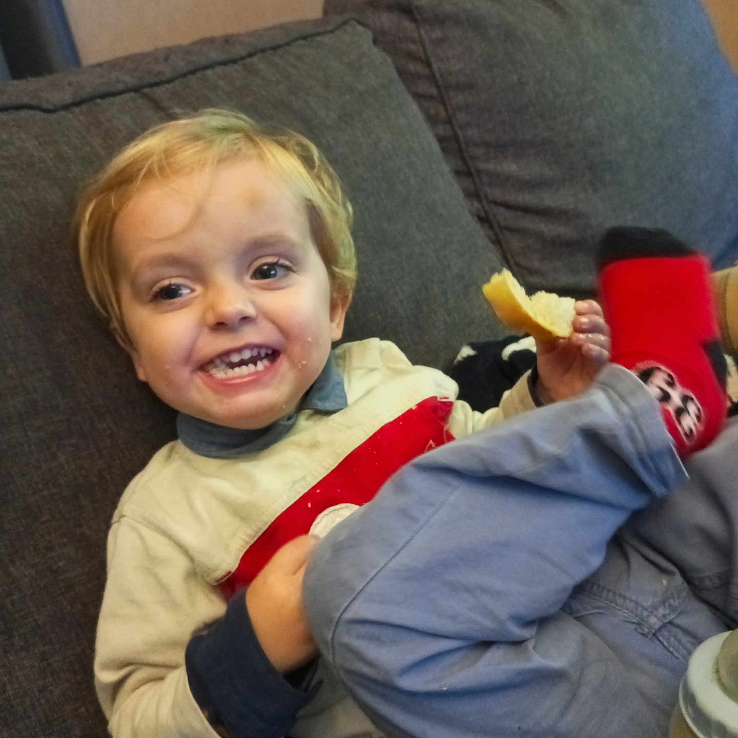 Maëlo participe au concours pour gagner de l'argent avec cette photo : blonde_hair, bread, casual_clothing, child, clothing, couch, cozy, face, food, fun, happy, home, indoor, person, red_socks, relaxed, sitting, smiling, toddler, young_child