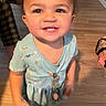 toddler, child, smile, indoor, floor, dress, patterned_clothing, happy, person, young_child, cute, joyful, standing, wooden_floor, holding_object, short_sleeves, curly_hair, playful, bright_eyes, casual