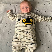 Vinnie is registered to the contest to win money with this photo: baby, infant, smile, onesie, clothing, blanket, stars, pattern, cute, happy, dimples, face, person, lying_down, child, spider, monster_design, text_on_clothing, indoors, soft_texture