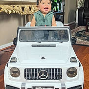 Josseling is registered to the contest to win money with this photo: toddler, toy_car, indoor, smiling, child, miniature_vehicle, wooden_floor, furniture, plant, happy, home, amg, car_grill, head, person, seat, window, greenery, clothing, living_room