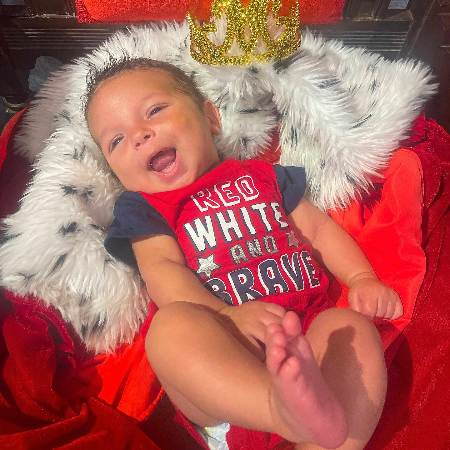 Oliver is registered to the contest to win money with this photo: baby, child, closeup, crown, cute, face, foot, hand, happy, indoors, infant, lying_down, orange_blanket, person, portrait, red_blanket, red_clothing, skin, smiling, white_fur