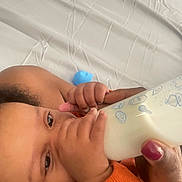 Leopold is registered to the contest to win money with this photo: baby, bottle, milk, feeding, hand, orange_clothing, infant, close_up, person, skin, face, eyes, nail_polish, bed, sheet, wrinkles, care, feeding_time, comfort, toddler