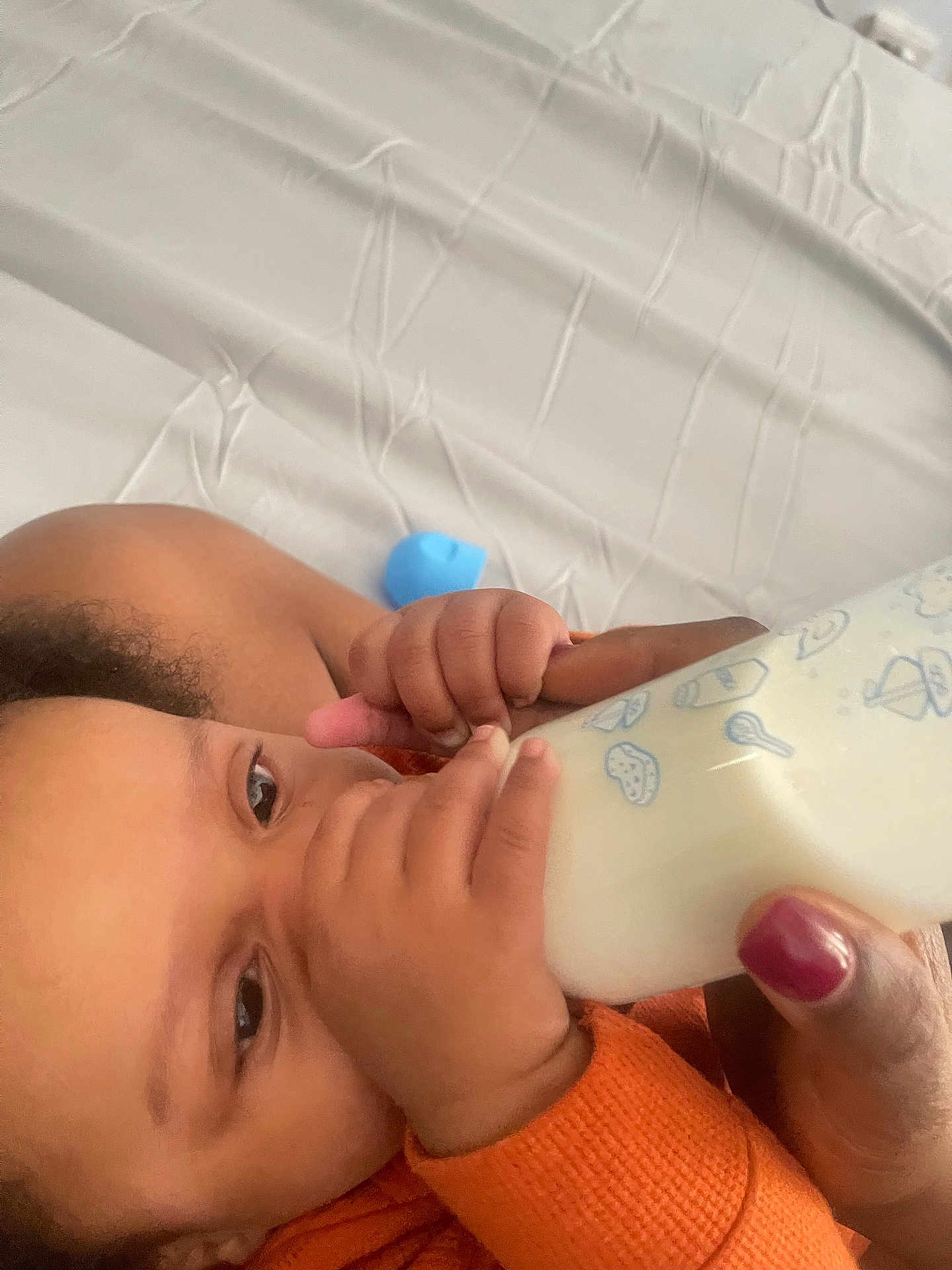 Leopold is registered to the contest to win money with this photo: baby, bottle, milk, feeding, hand, orange_clothing, infant, close_up, person, skin, face, eyes, nail_polish, bed, sheet, wrinkles, care, feeding_time, comfort, toddler