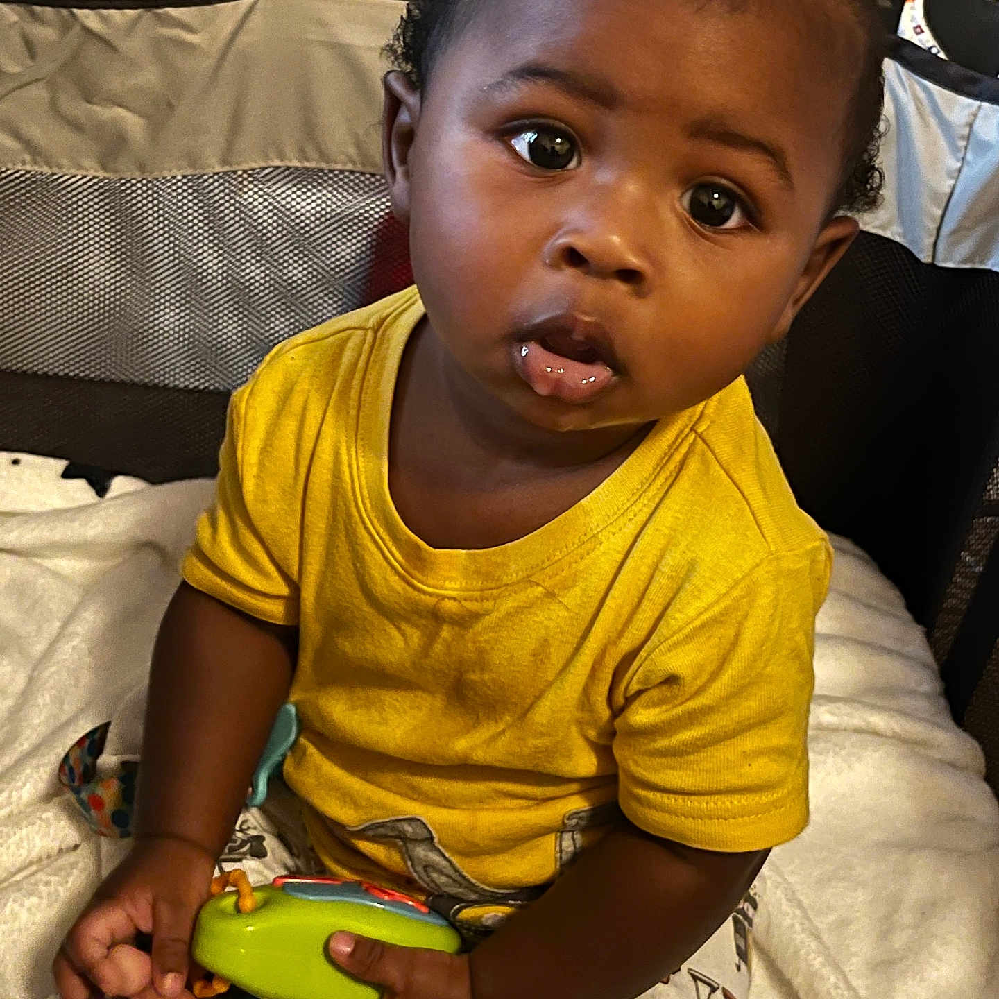 Kevion is registered to the contest to win money with this photo: baby, blanket, bright_colors, child, clothing, curious, cute, expression, foot, hand, indoor, infant, person, playpen, portrait, sitting, soft_texture, toy, yellow_shirt, young_child