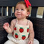 toddler, child, smile, red_bow, strawberry_pattern, crochet_top, white_pants, wicker_chair, outdoor, tree, wooden_fence, cute, portrait, baby, sitting, happy, earrings, short_hair, natural_light, casual_clothing