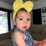 Jaylene is registered to the contest to win money with this photo: toddler, child, yellow_bow, headband, floral_dress, indoor, couch, living_room, young_child, cute, portrait, person, face, skin, eyes, hair, earrings, furniture, window, home