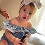 Jaylene is registered to the contest to win money with this photo: baby, child, infant, headband, bow, remote_control, blue_clothing, off_shoulder, sitting, indoor, window, blinds, hand, curious, cute, face, portrait, light, skin, table