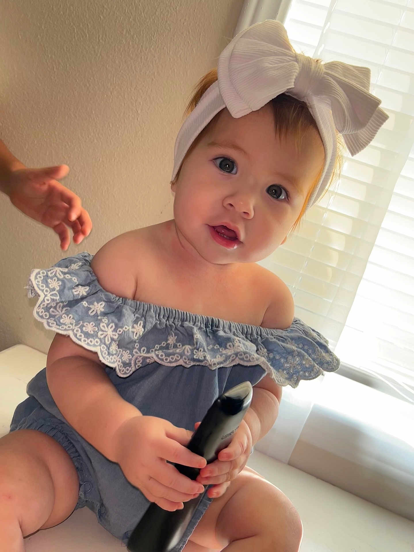Jaylene is registered to the contest to win money with this photo: baby, child, infant, headband, bow, remote_control, blue_clothing, off_shoulder, sitting, indoor, window, blinds, hand, curious, cute, face, portrait, light, skin, table