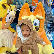 Malachi joined the competition — help win amazing prizes! baby, child, plush_toy, stuffed_animal, shopping_cart, bear_onesie, orange_plush, yellow_plush, cute, wide_eyes, hand, clothing, indoors, toy_store, soft_toy, person, animal_costume, cart, expression, seated