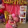 baby, birthday, pink_dress, crown, balloon, carpet, smiling, decorations, gold_balloon, number_one, stacked_boxes, party, celebration, indoor, happy, child, festive, colorful, toddler, cute