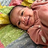 apparel, baby, blanket, child, closeup, colorful, cute, expression, face, happy, indoor, infant, lying_down, pink_clothing, portrait, quilt, reaching_hand, skin, smile, young