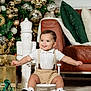 baby, child, smiling, tongue_out, chair, white_shoes, suspenders, shorts, christmas_tree, ornaments, presents, carpet, couch, pillows, holiday, indoor, decorations, festive, happy, portrait