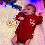 baby, infant, red_onesie, blanket, wooden_sign, text, face, legs, arms, christmas_tree_branch, blue_light, holiday, cozy, newborn, cute, lying_down, indoor, soft_texture, celebration, portrait