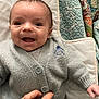 baby, smiling, cardigan, gray_clothing, hand, quilted_blanket, colorful_blanket, infant, happy, closeup, face, portrait, cute, cozy, lying_down, button, human_hand, warm, soft_texture, indoor